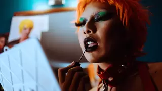 Non-binary person with orange hair applies makeup in a mirror